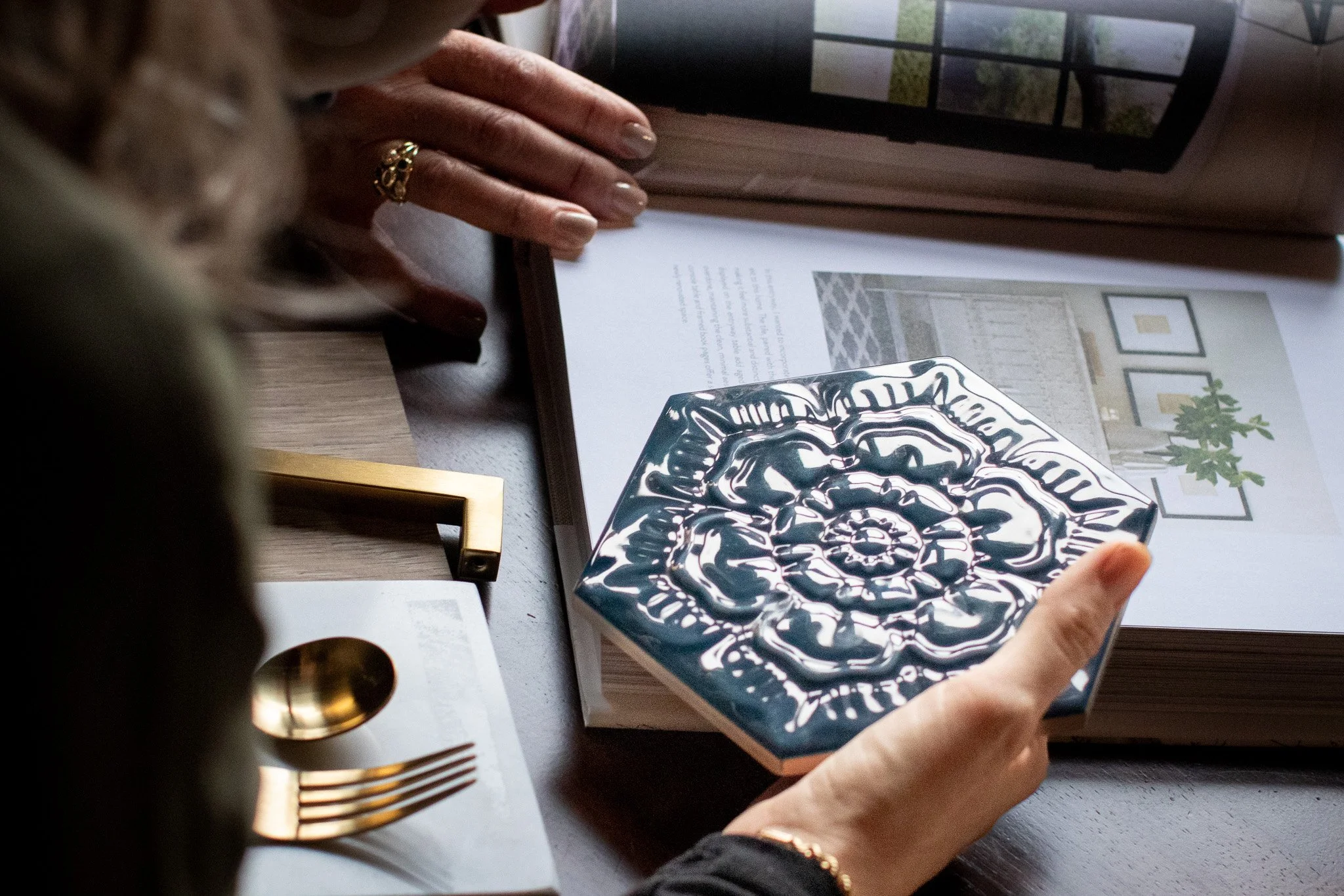Amber Parker holding a decorative ceramic tile at a table with an open magazine, gold cutlery, and a framed photo at an interior design client meeting. .