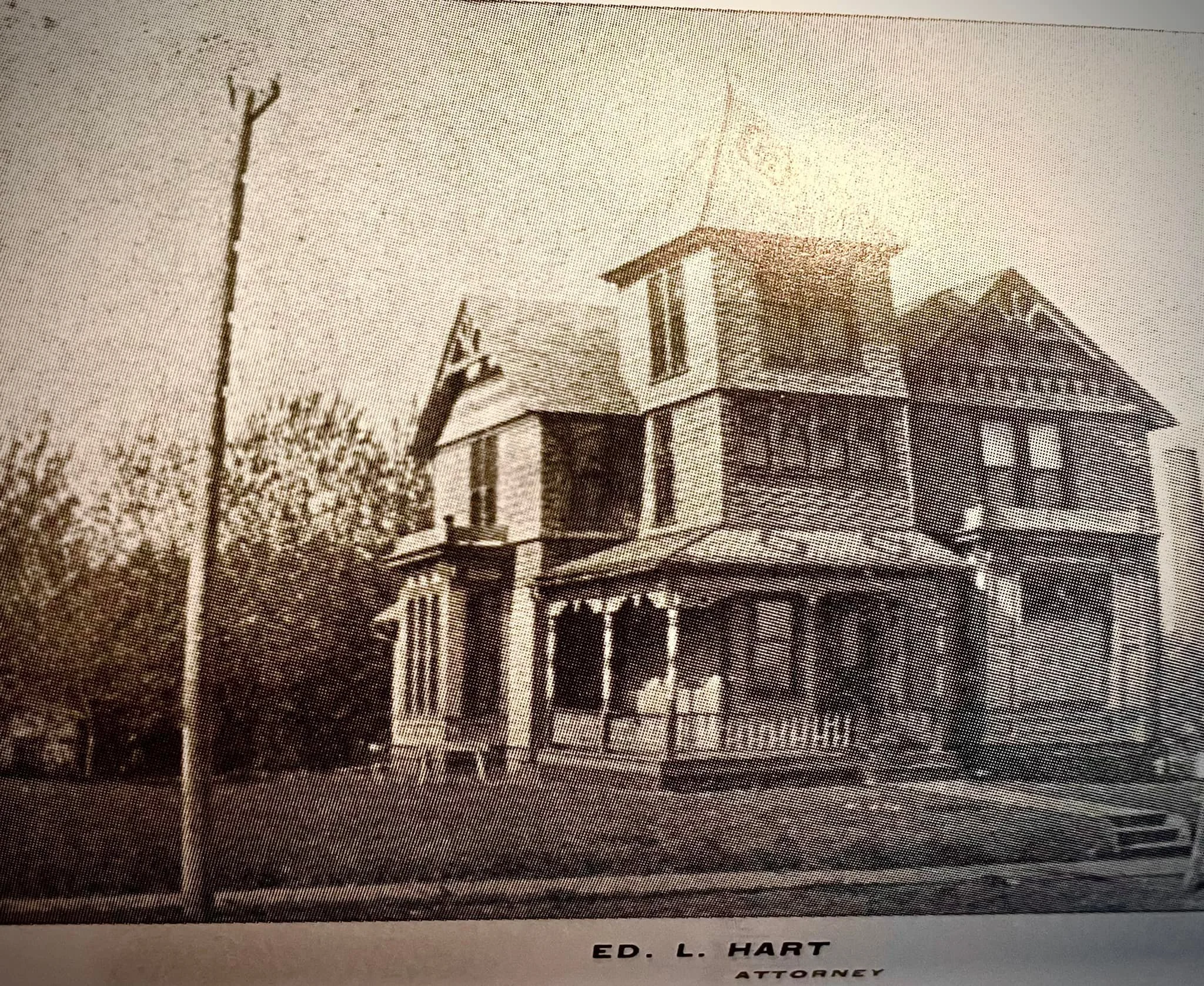 A black and white photo of a two-story Victorian-style house with a porch, located in a neighborhood, with a flagpole nearby and some trees in the background. The caption at the bottom reads "ED. L. HART ATTORNEY."