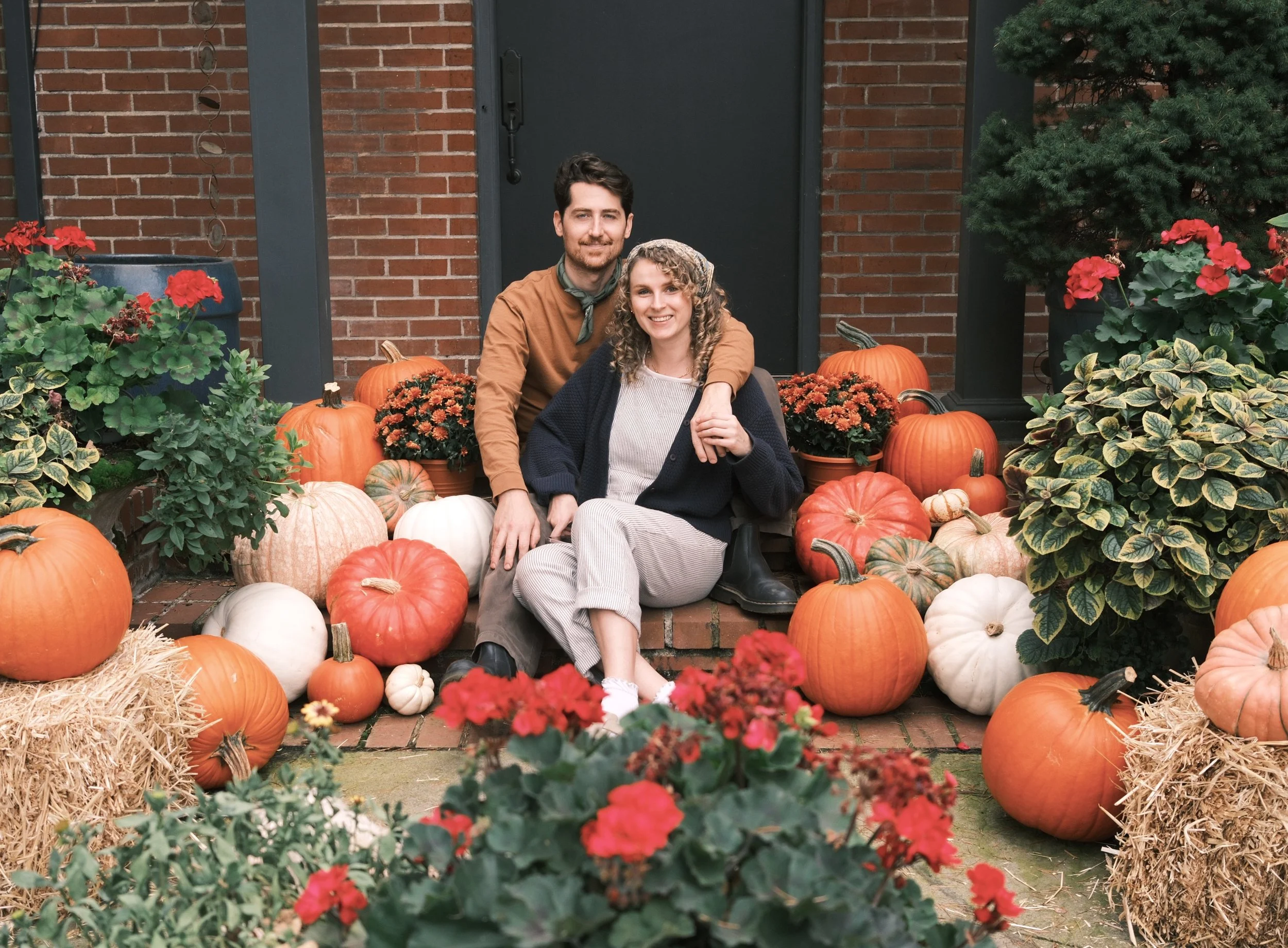 A smiling man with a beard and a woman with curly hair sit together on a step in front of a beautiful brick home. They are surrounded by an assortment of pumpkins, gourds, hay bales, and potted flowers, indicating a fall or harvest season setting.