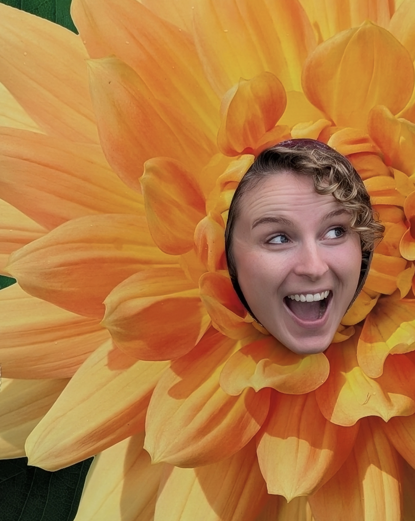 A woman with curly hair smiling and making a surprised face, with her face through a circular cutout in the center of a large orange and yellow flower.
