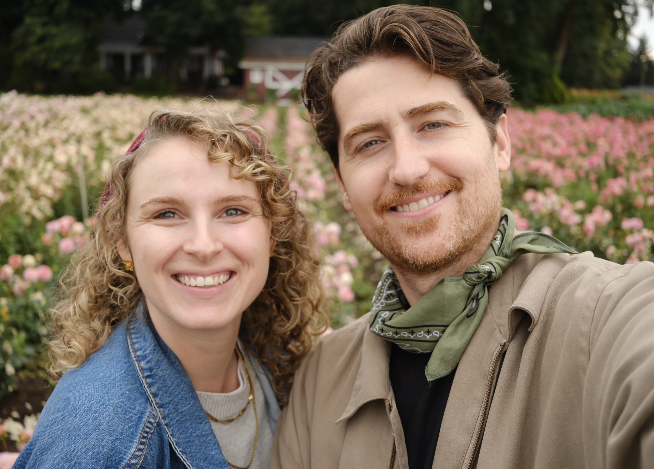 A young woman and a man smiling as they take a selfie outdoors in a garden with pink flowers and trees.