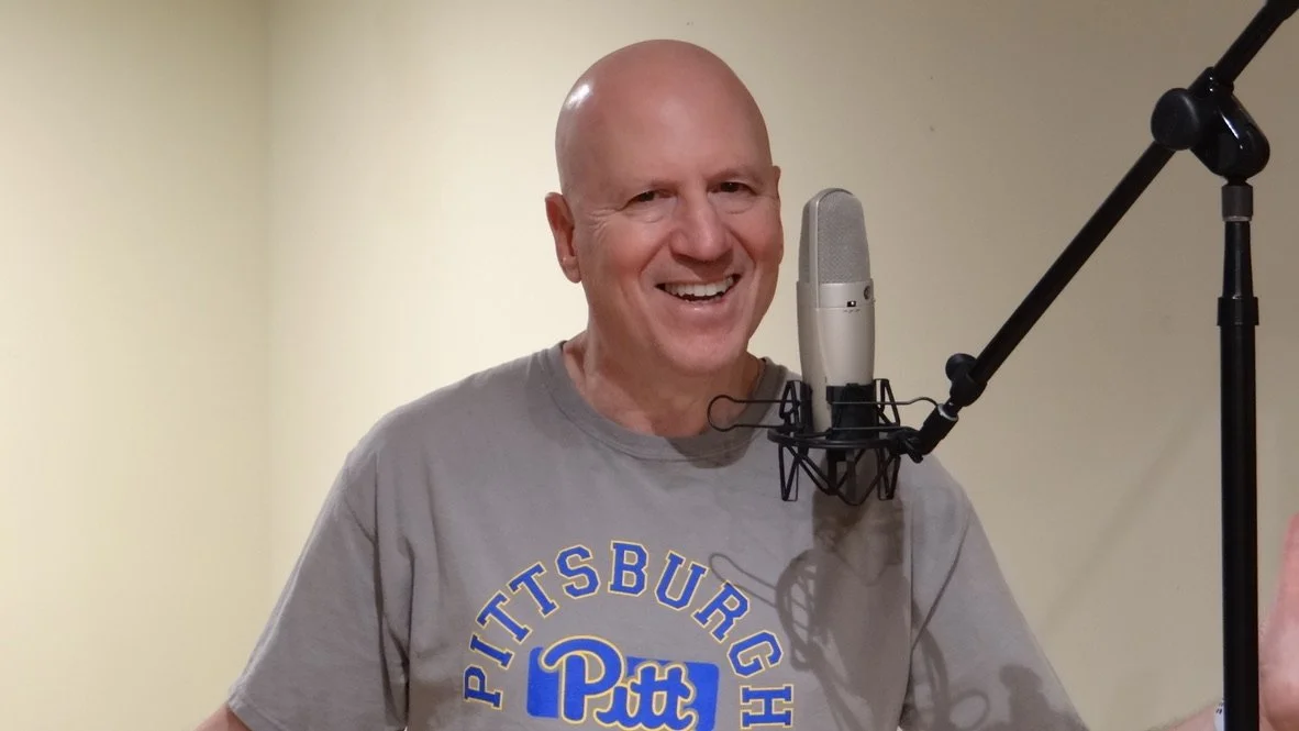 Dylan Jordan - A smiling man standing in front of a microphone with a pop filter, wearing a gray Pitt Panthers t-shirt, in a recording studio or room.