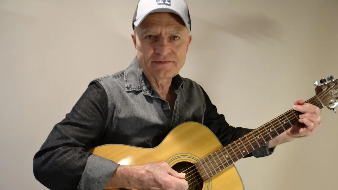 Phil Barrile - An older man wearing a white cap and a black and gray shirt playing an acoustic guitar.