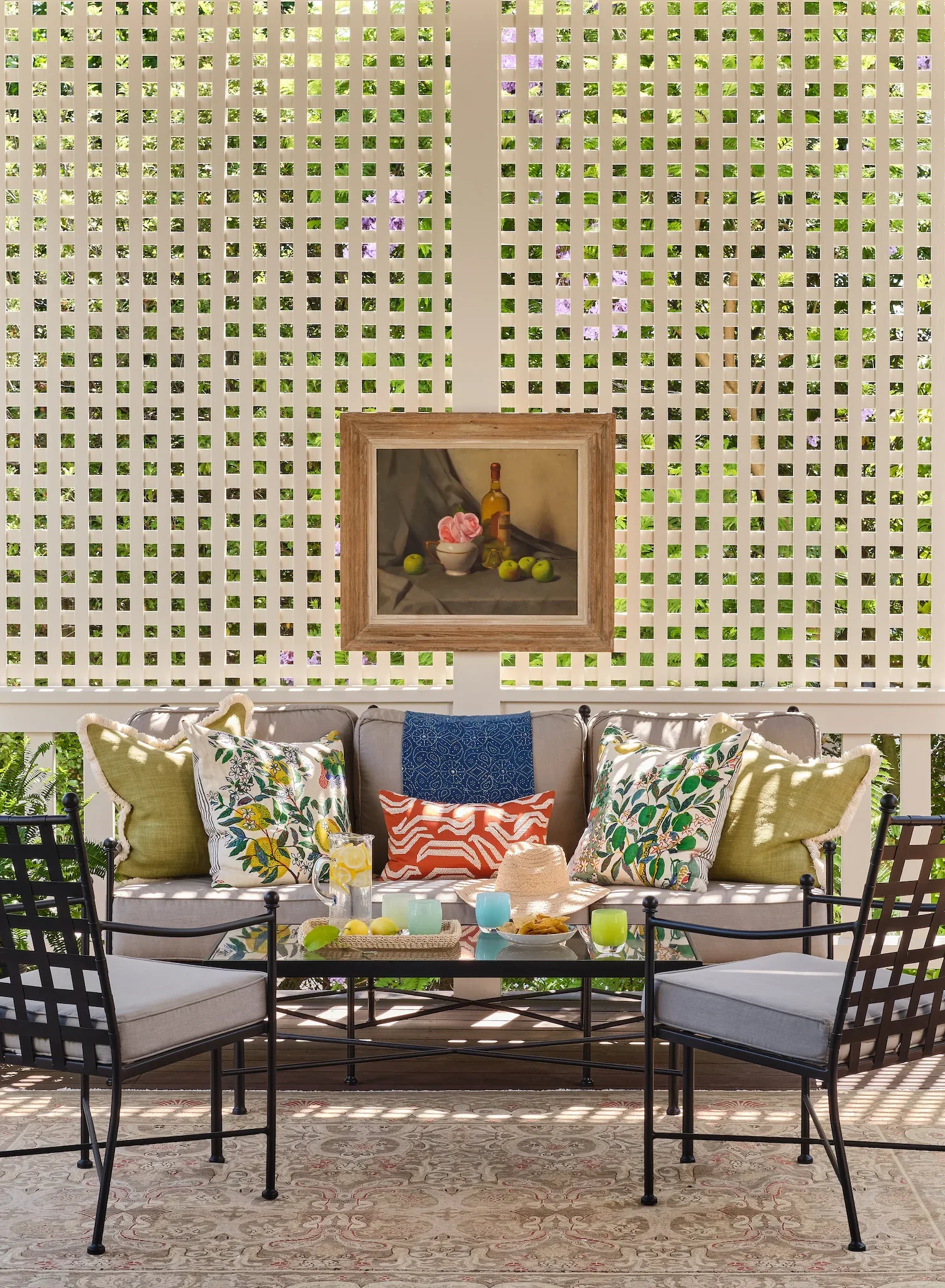 A cozy outdoor seating area with a white cushioned sofa decorated with various colorful floral and geometric patterned pillows. In front of the sofa, there is a black metal table with glasses, a pitcher of lemon water, and snacks. Behind the sofa, a white lattice fence allows greenery and purple flowers to show through. A framed still-life painting of a bottle, bowl of roses, apples, and a glass adorns the fence. The area has a warm, inviting atmosphere with natural sunlight illuminating the space.