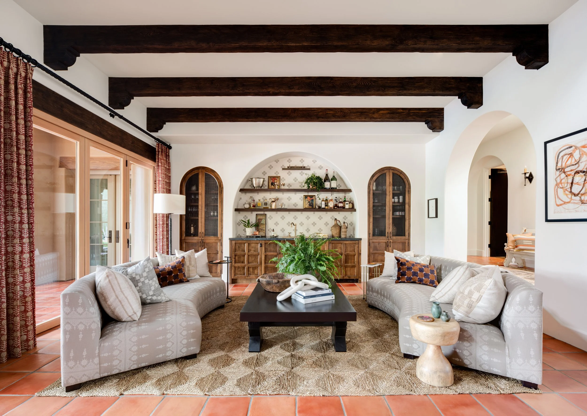 Living room with two white sofas with patterned pillows, a black coffee table, potted plant, and decorative items. There are wood cabinets and shelves in the background, and terracotta tiled flooring.