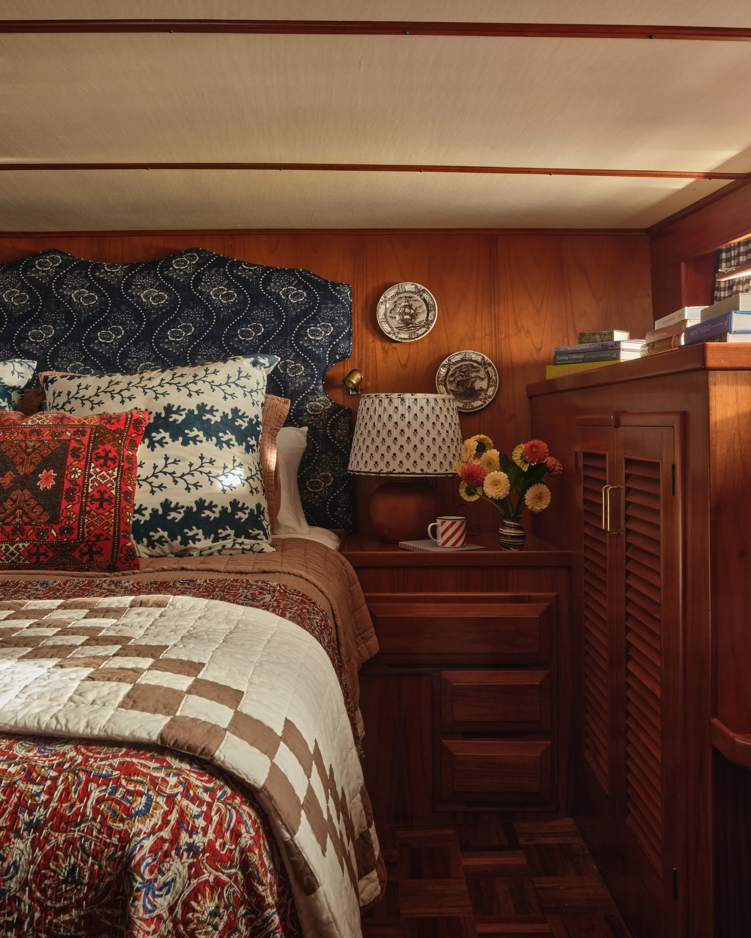 Boat stateroom with wooden walls and trim, featuring bedding with a checkered pattern, decorative pillows, hanging decorative plates, a bedside table with a lamp, books, a vase of flowers, and a partially open louvered wooden cabinet.