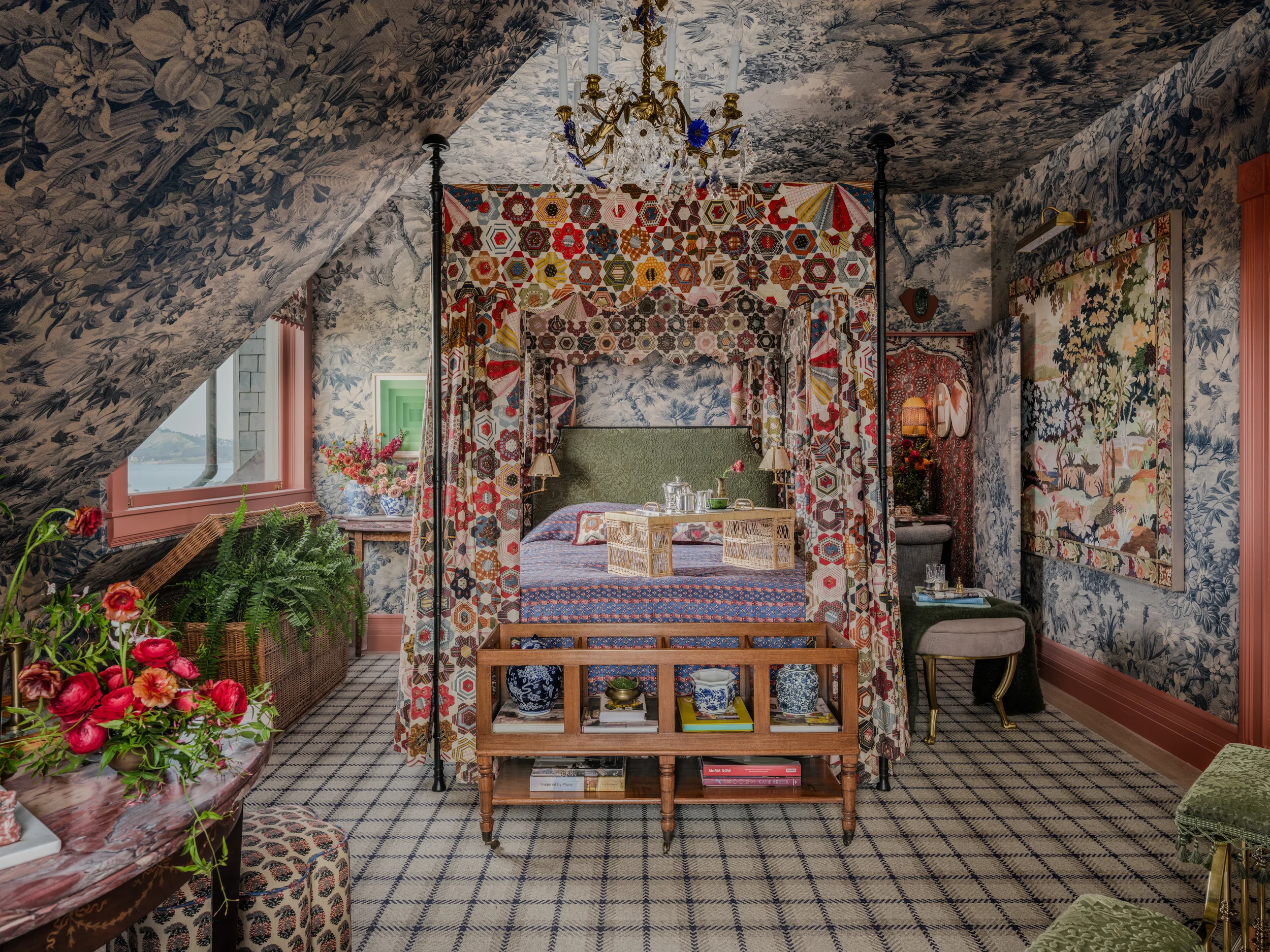 A cozy vintage-style bedroom with floral and geometric patterned wallpaper and curtains, featuring a bed with a green headboard, a quilted canopy, a dresser with vases and flowers, and a chandelier hanging from the ceiling.