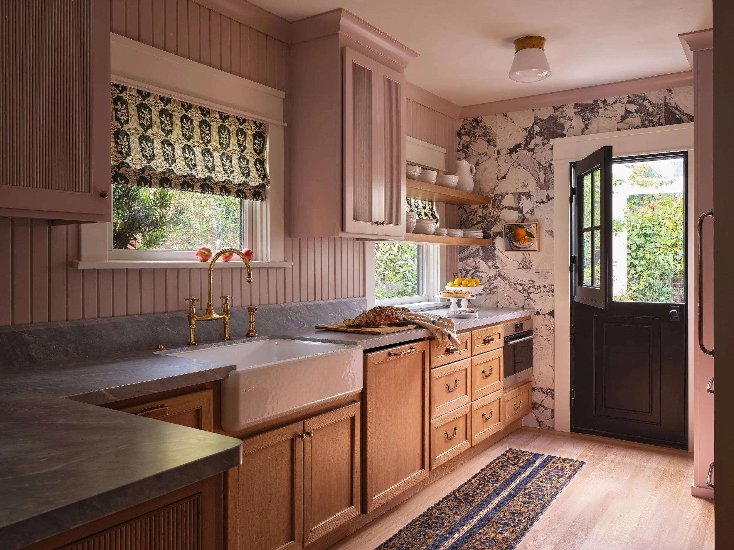 A kitchen with light wooden cabinets, a marble countertop, a farmhouse sink with a brass faucet, open shelves with dishes, a window with a patterned curtain, and a door leading outside with greenery visible.