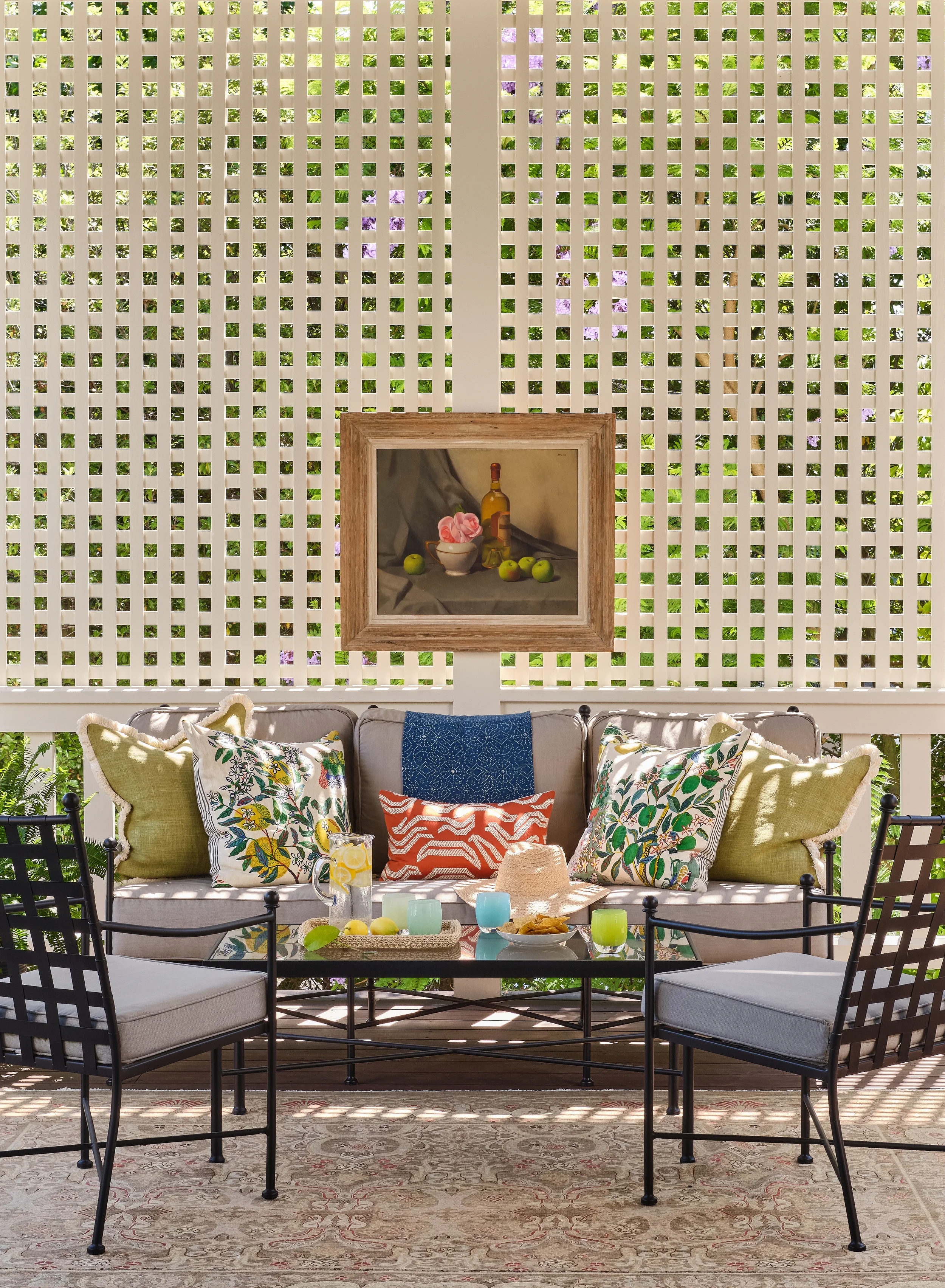 Outdoor patio seating area with gray cushions, colorful pillows, a glass table with drinks, snacks, and a straw hat, surrounded by black metal chairs, against a white lattice wall with plants and purple flowers behind it.
