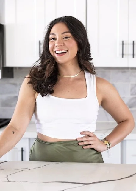 A young woman with dark hair smiling in a kitchen, wearing a white sleeveless top and green pants.