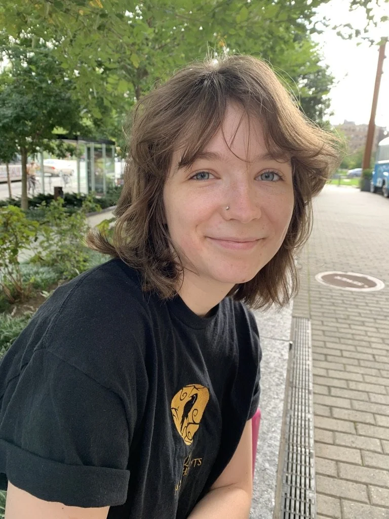 Young woman with wavy brown hair, wearing a black T-shirt with a yellow and black logo, sitting outdoors with green trees and sidewalk in the background, smiling softly at the camera.