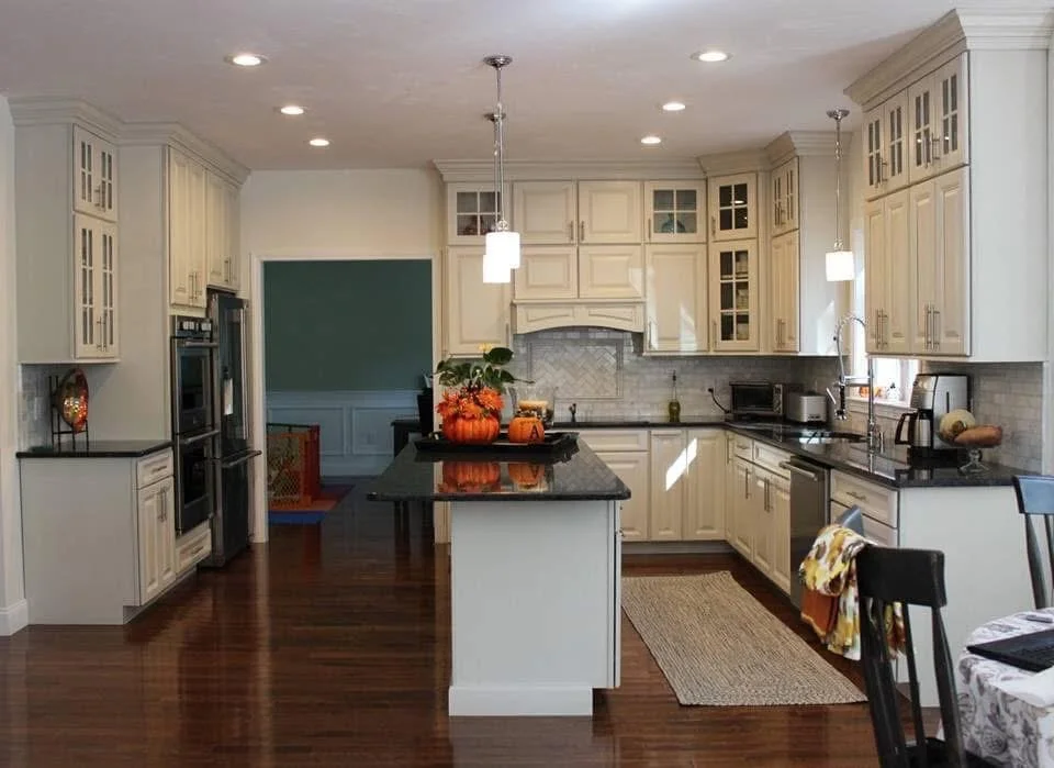 Bright kitchen with white cabinets, black countertops, hardwood floors, and modern pendant lighting. Decorated with fall or Halloween themed items, including pumpkins and a bouquet, on the central island.