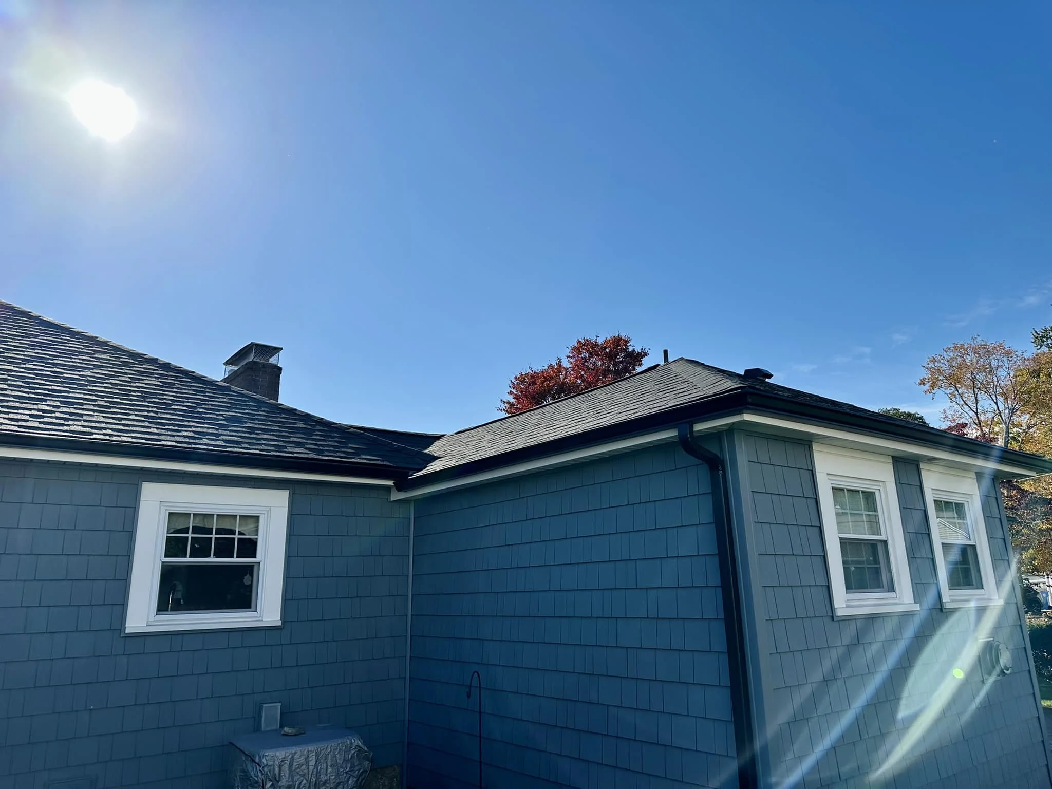 Blue house with white window frames, roof with asphalt shingles, sun shining with lens flare, trees with autumn foliage in the background.