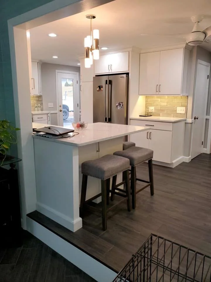 Modern white kitchen with a small breakfast bar, two gray stools, stainless steel refrigerator, and pendant lighting.