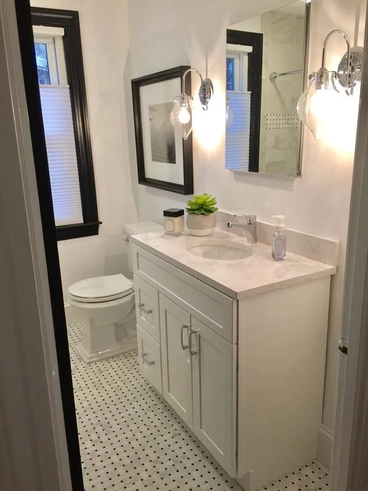 Small bathroom with white vanity, marble countertop, round sink, soap dispenser, potted plant, framed art, two small windows with black trim, and wall-mounted light fixtures.