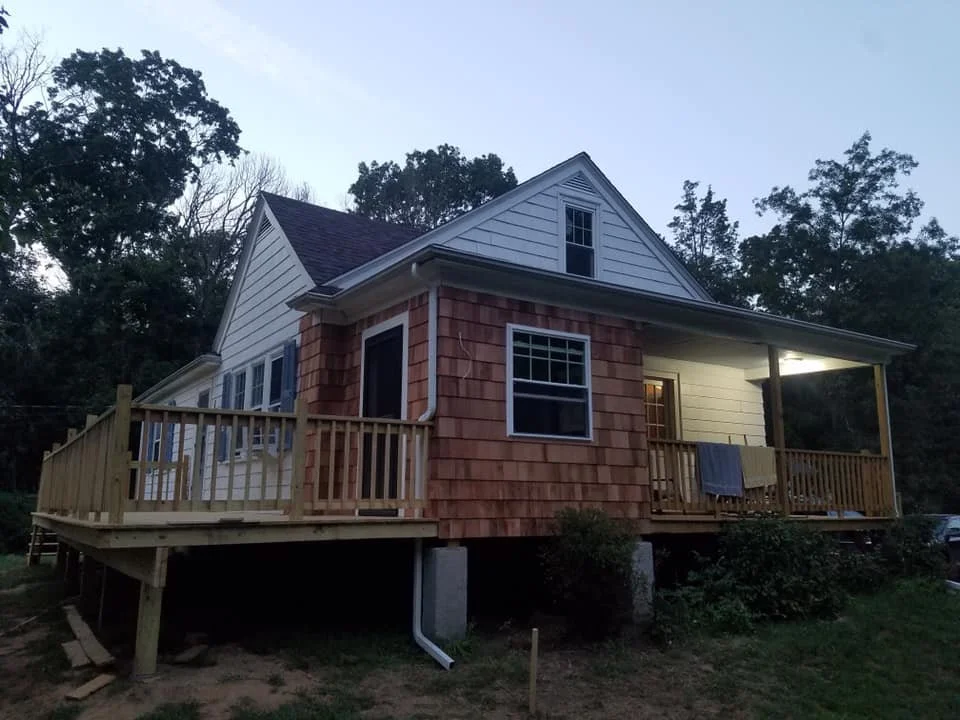 A two-story house under construction with a large front porch, surrounded by trees.