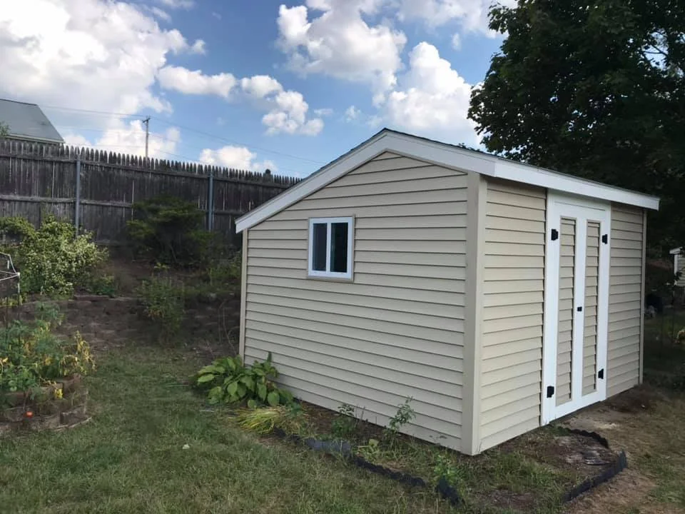 A beige garden shed with double doors and a small window, situated in a backyard with a wooden fence, plants, and a cloudy sky.