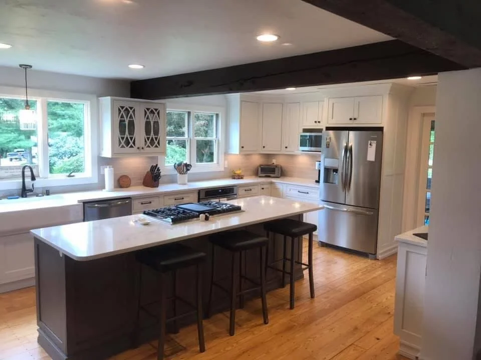 Modern kitchen with white cabinets, stainless steel refrigerator, microwave, countertop oven, and a kitchen island with stools, hardwood floors, large windows with greenery outside, and recessed ceiling lights.