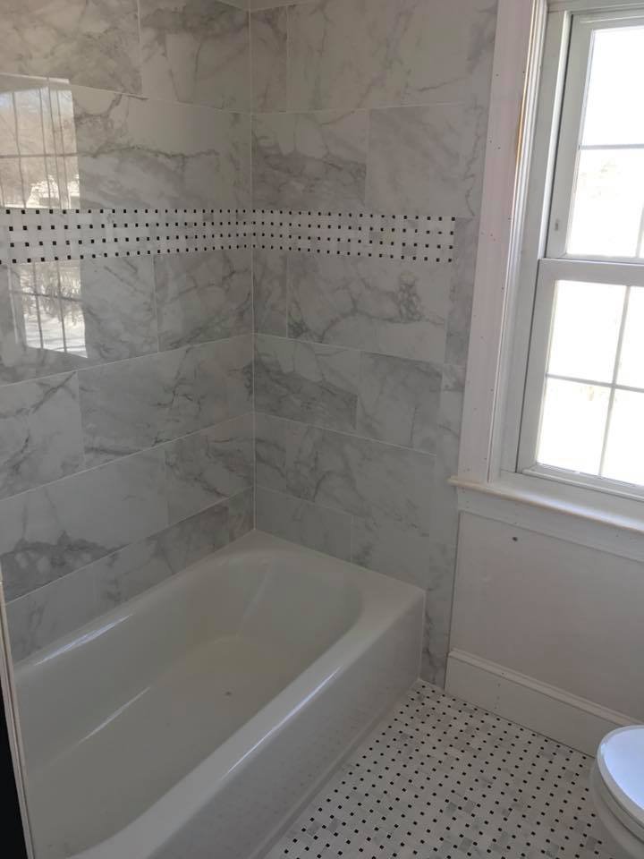 Bathroom with a white bathtub, marble wall tiles, a window with white trim, and small black and white mosaic floor tiles.