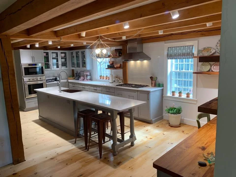 Modern kitchen with grey cabinetry, white countertops, and a kitchen island, featuring wooden ceiling beams, a stainless steel range hood, potted plants, and open shelves with decorative bowls.