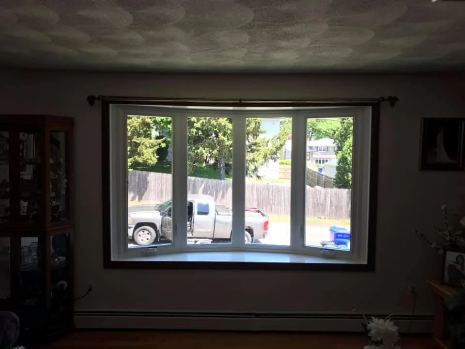 View from inside a room looking out a large window to a suburban street with a pickup truck parked outside, a wooden fence, trees, and a house in the background.