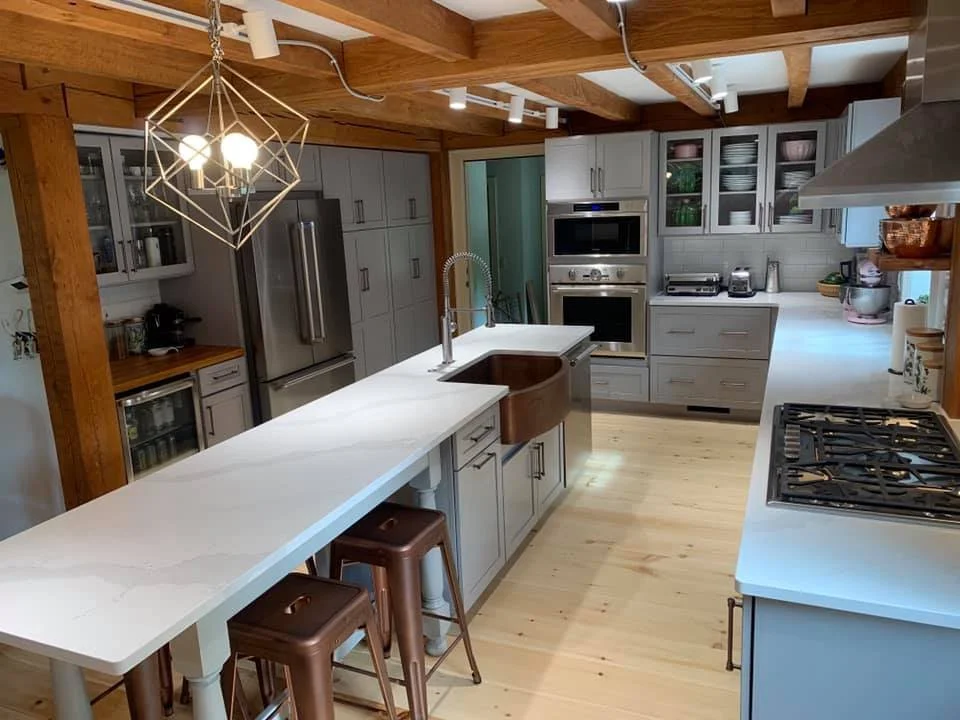 Modern kitchen with gray cabinets, white countertops, a wooden ceiling with exposed beams, and a central island with a rust-colored sink. There's a gas stove, built-in oven, and stainless steel refrigerator. Wooden stools sit at the counter, and a ge