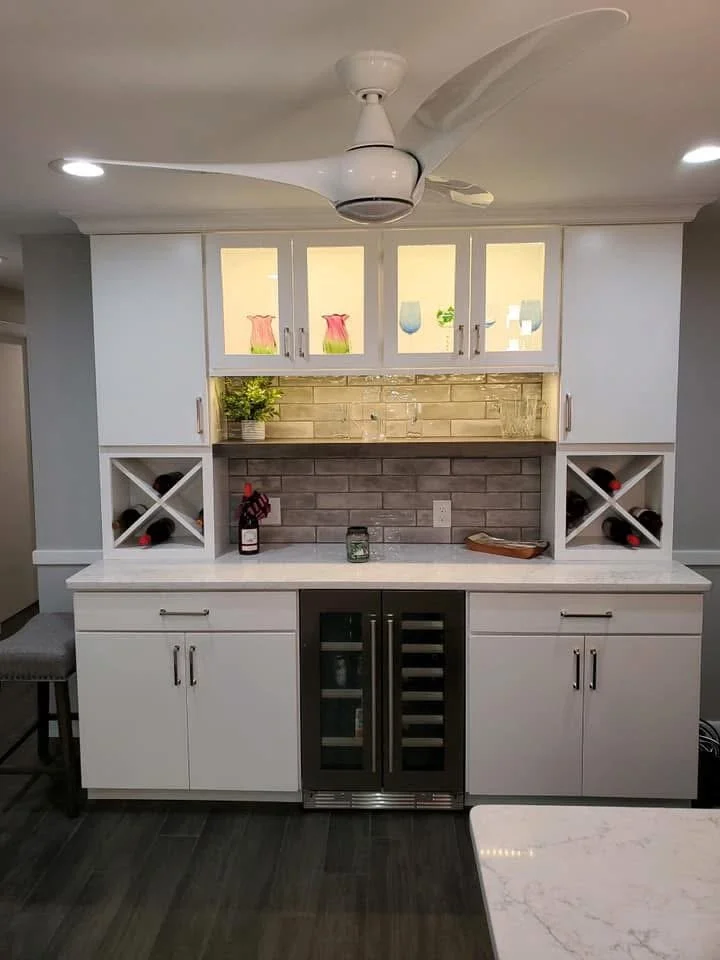 White kitchen bar area with wine storage, a mini wine fridge, and upper cabinets with decorative vases and glasses, under a ceiling fan.