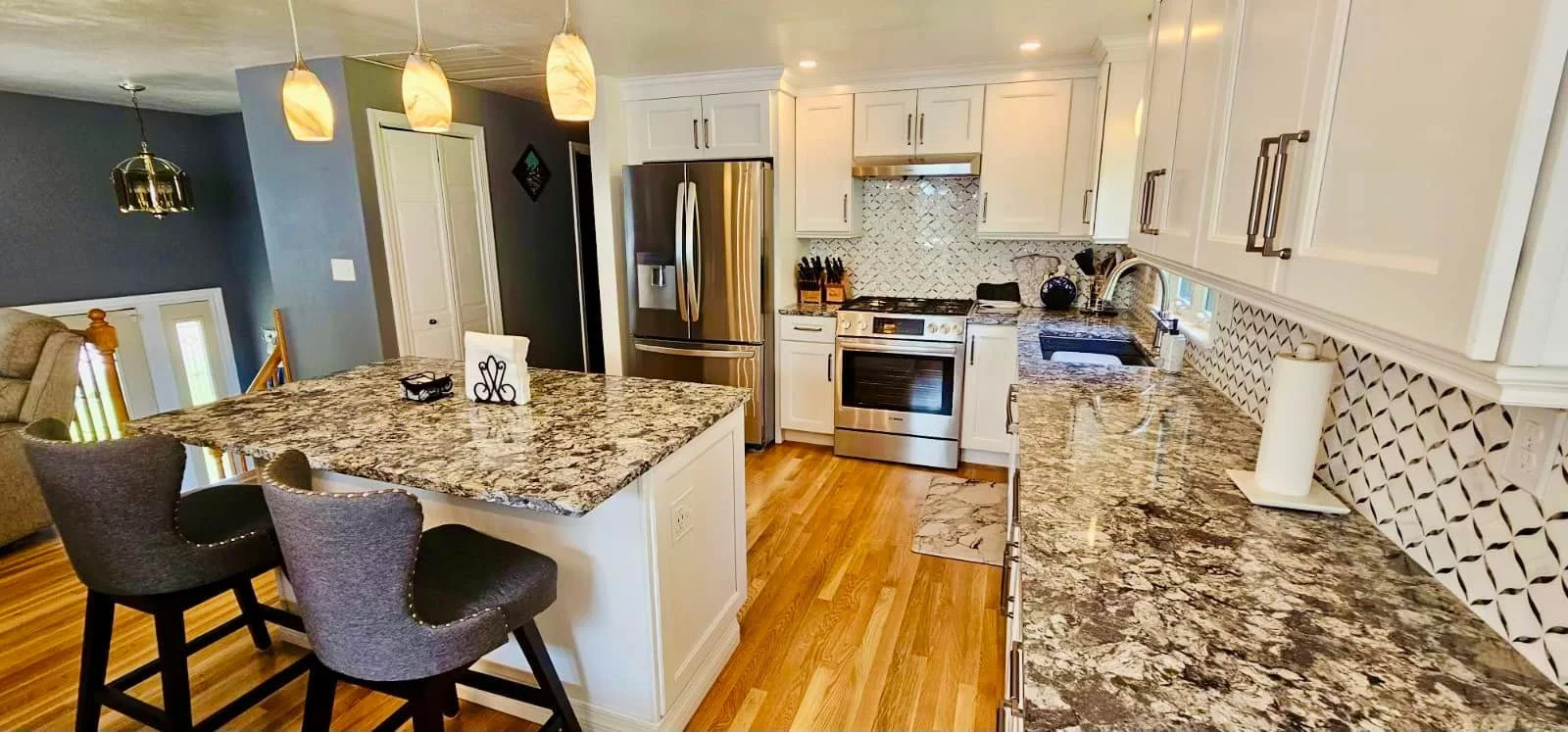 Modern kitchen with granite countertops, white cabinets, stainless steel refrigerator, stove, and black sink. There are three pendant lights hanging over the island with two grey chairs.
