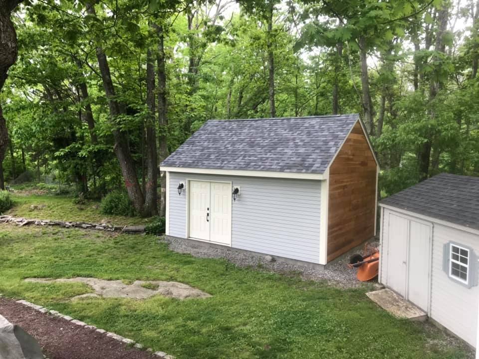 A small, newly built shed with light gray siding, white doors, black outdoor lights, and a gray shingled roof, situated on a grassy lawn with trees in the background.