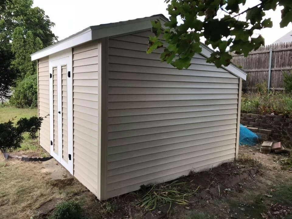 A beige plastic shed with white double doors, located on a grassy backyard with trees and a wooden fence.
