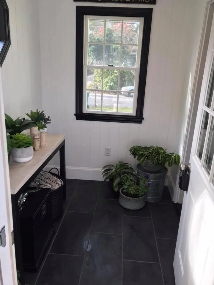 Sunlit indoor room with black-framed window, white walls, black tiled floor, potted plants on the right, and a shelf with decorative items on the left.