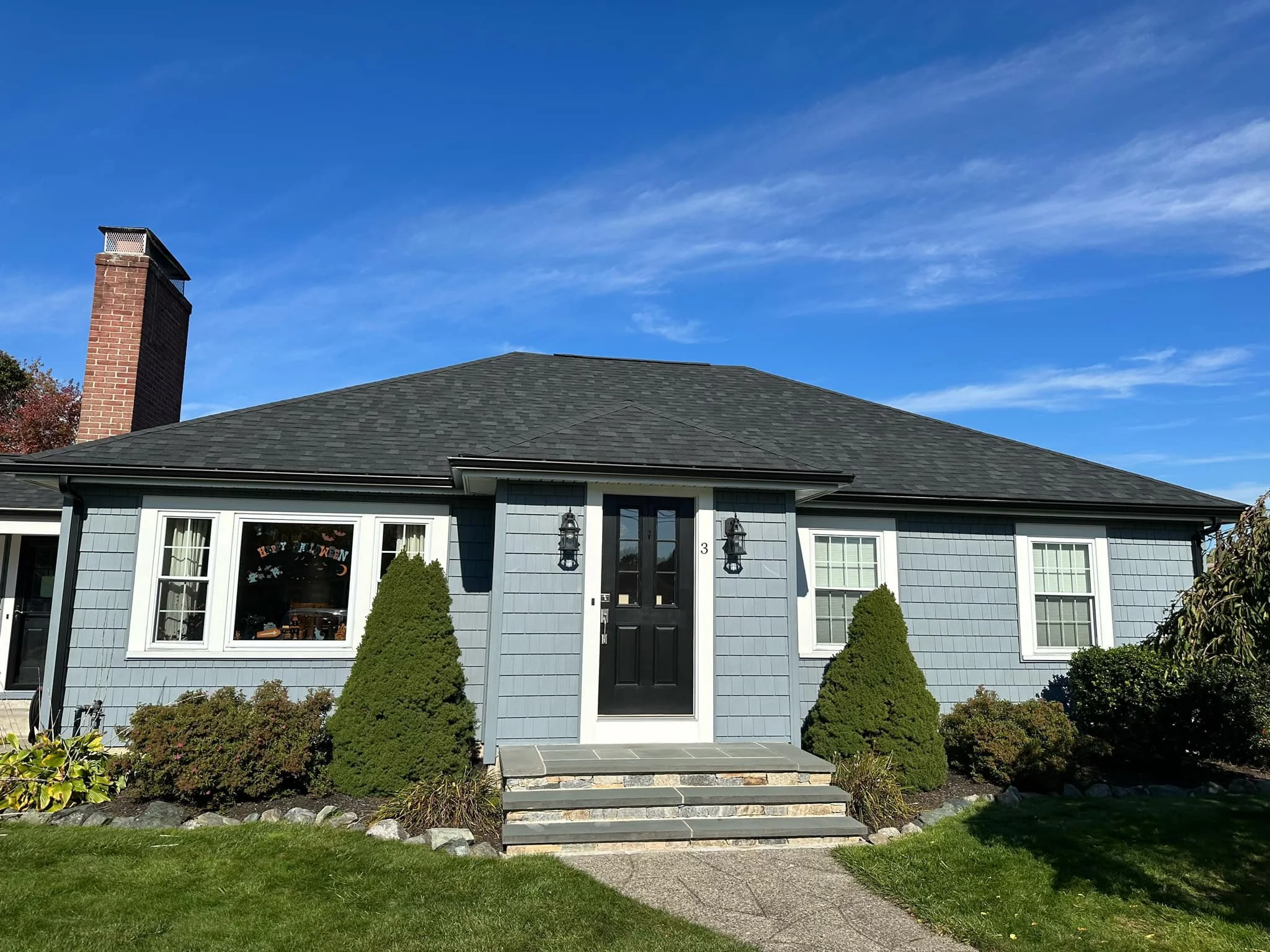 A house with light blue siding, three front steps leading to a black door, two large bushes flanking the door, white-framed windows, and a porch light on either side of the door, under a clear blue sky.