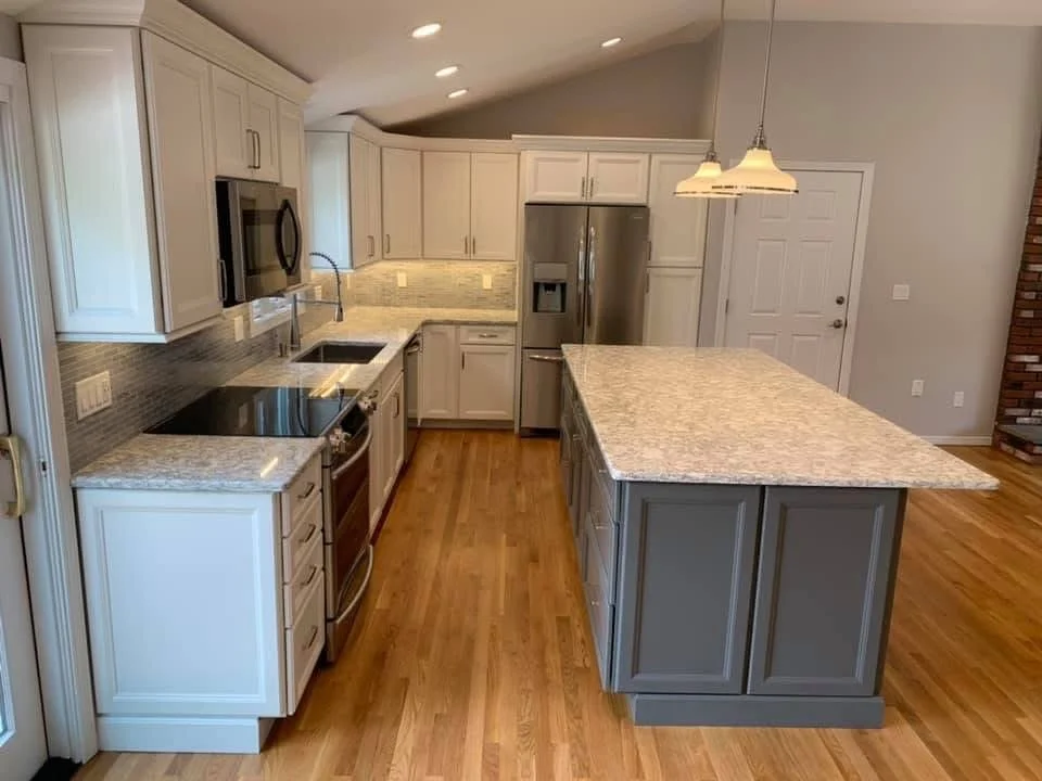 A modern kitchen with white cabinets, granite countertops, stainless steel appliances, and a wooden floor. There is an island with gray cabinetry, a hanging light fixture, and a door leading outside or to another room.