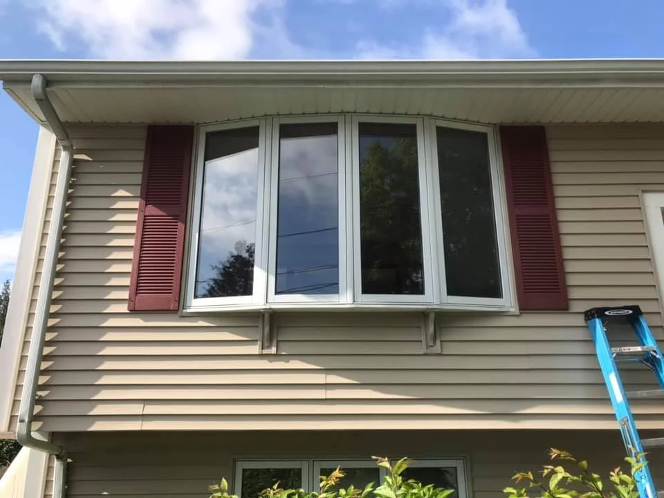 The image shows the exterior of a house with beige siding, a large curved window with white trim, and red shutters on either side. There is a blue ladder leaning against the house on the right side, and some green foliage at the bottom.