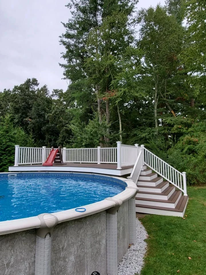 Above-ground swimming pool with staircase and white safety railing, surrounded by green grass, trees, and a small children's slide in the backyard.