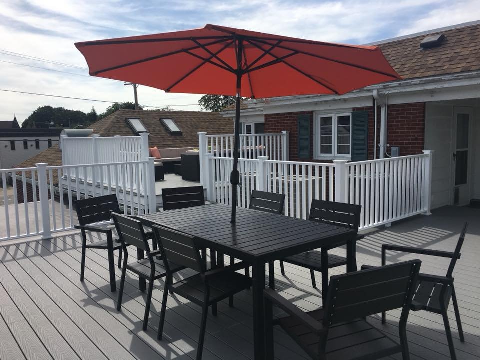 An outdoor rooftop deck with a black dining table, six matching chairs, and a large red umbrella, overlooking neighboring rooftops and partly cloudy sky.