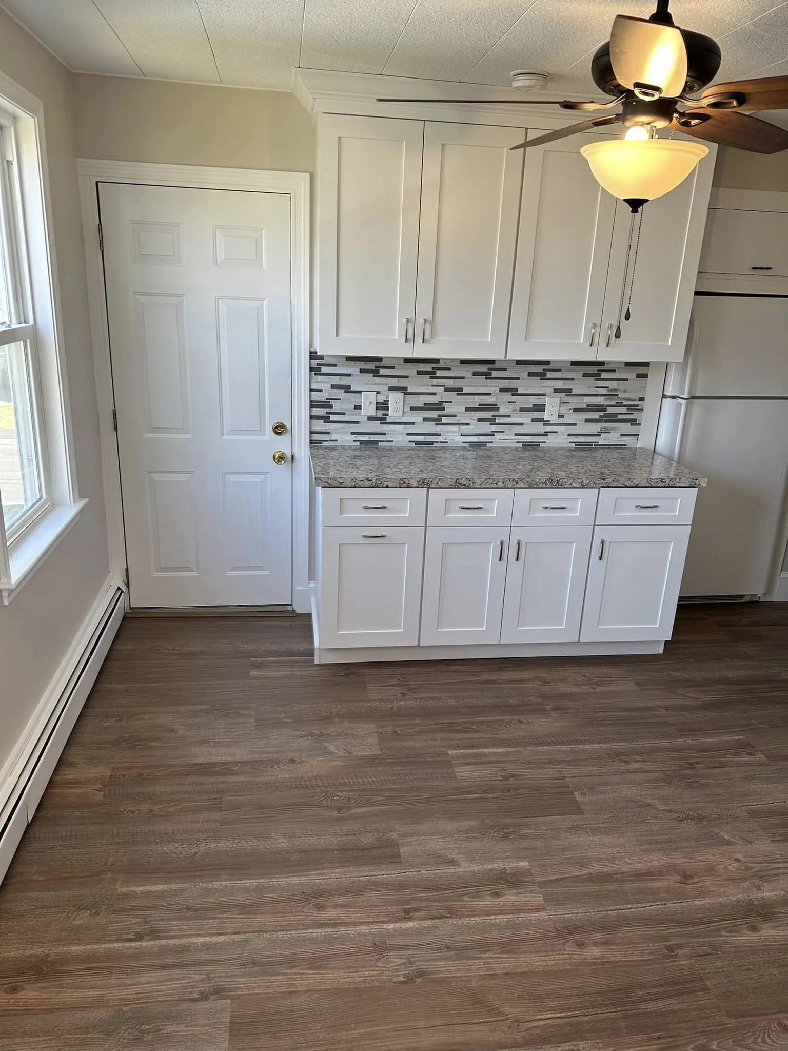Kitchen with white cabinets, granite countertop, and a grey, black, and white mosaic tile backsplash. Wooden flooring and a ceiling fan with a light are visible.