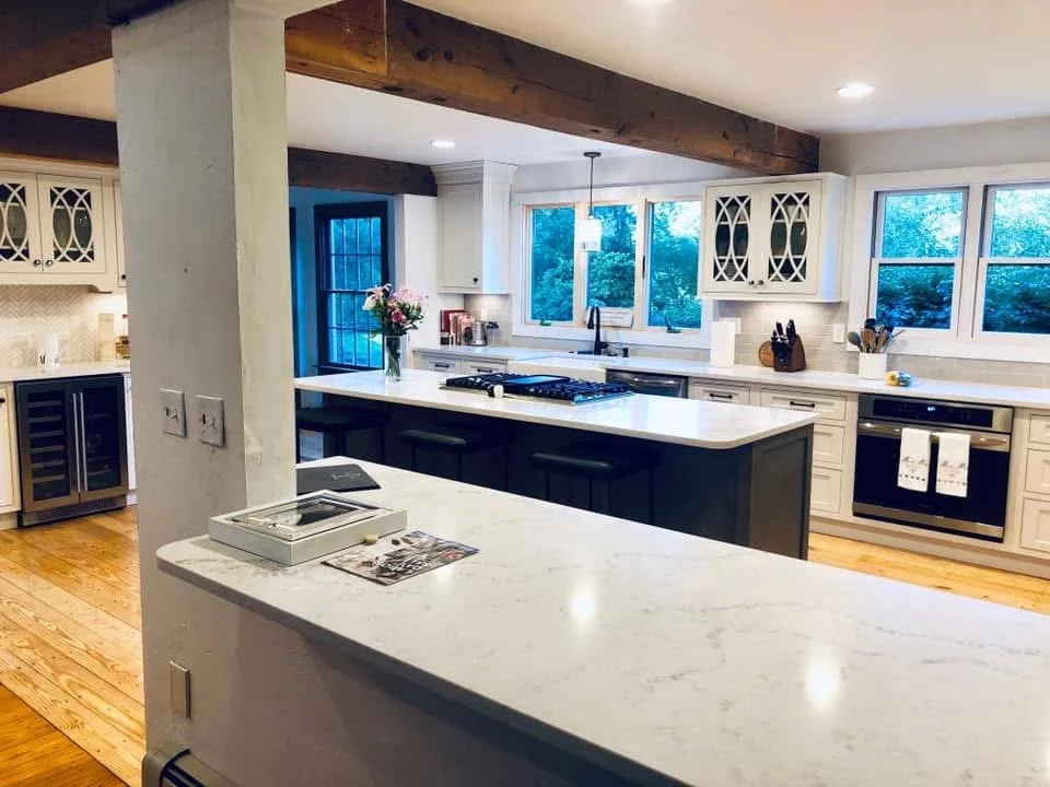 Modern kitchen with white cabinets, a black island, and large windows overlooking trees, with wooden ceiling beams and hardwood floors.