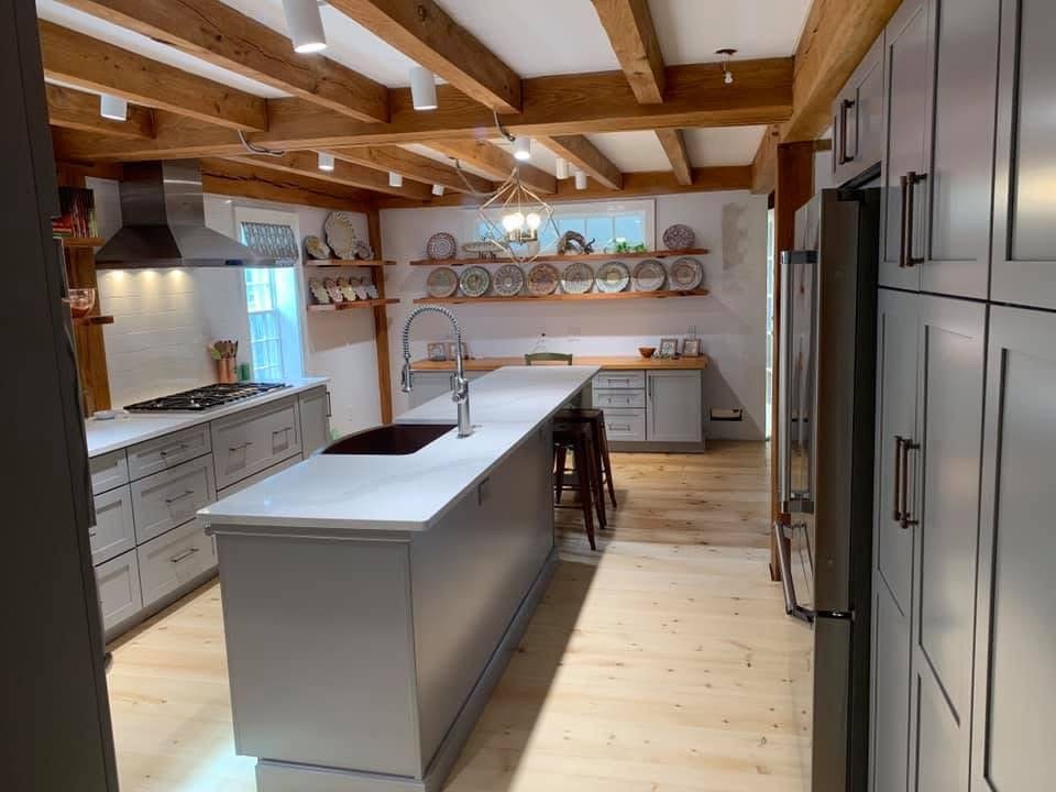 Modern kitchen with wooden beams, white cabinets, an island with a sink, open shelving with plates, and a window with natural light.