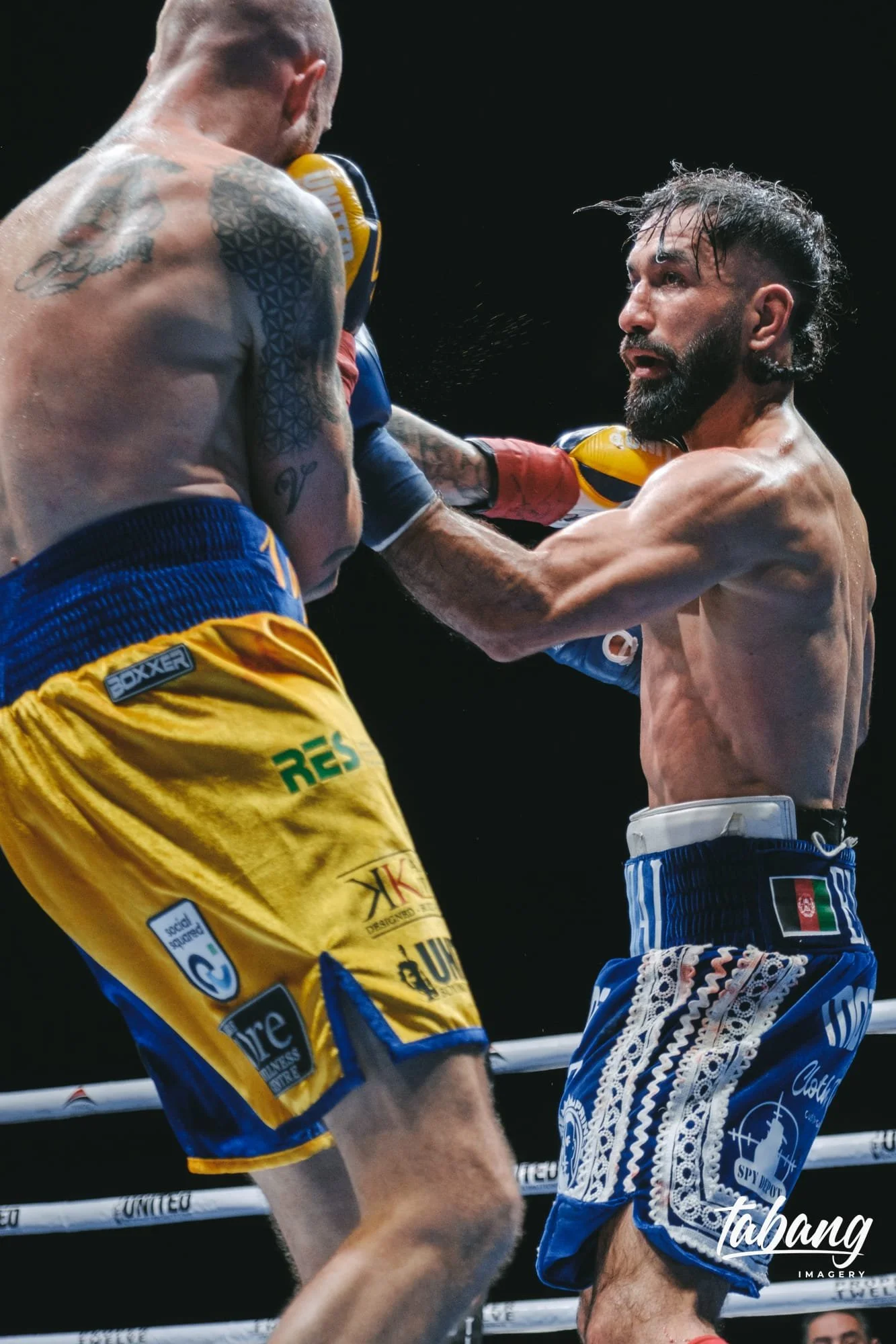 Two male fighters in a boxing match. One fighter with tattoos and yellow shorts, the other with a beard and blue shorts, being hit in the face.