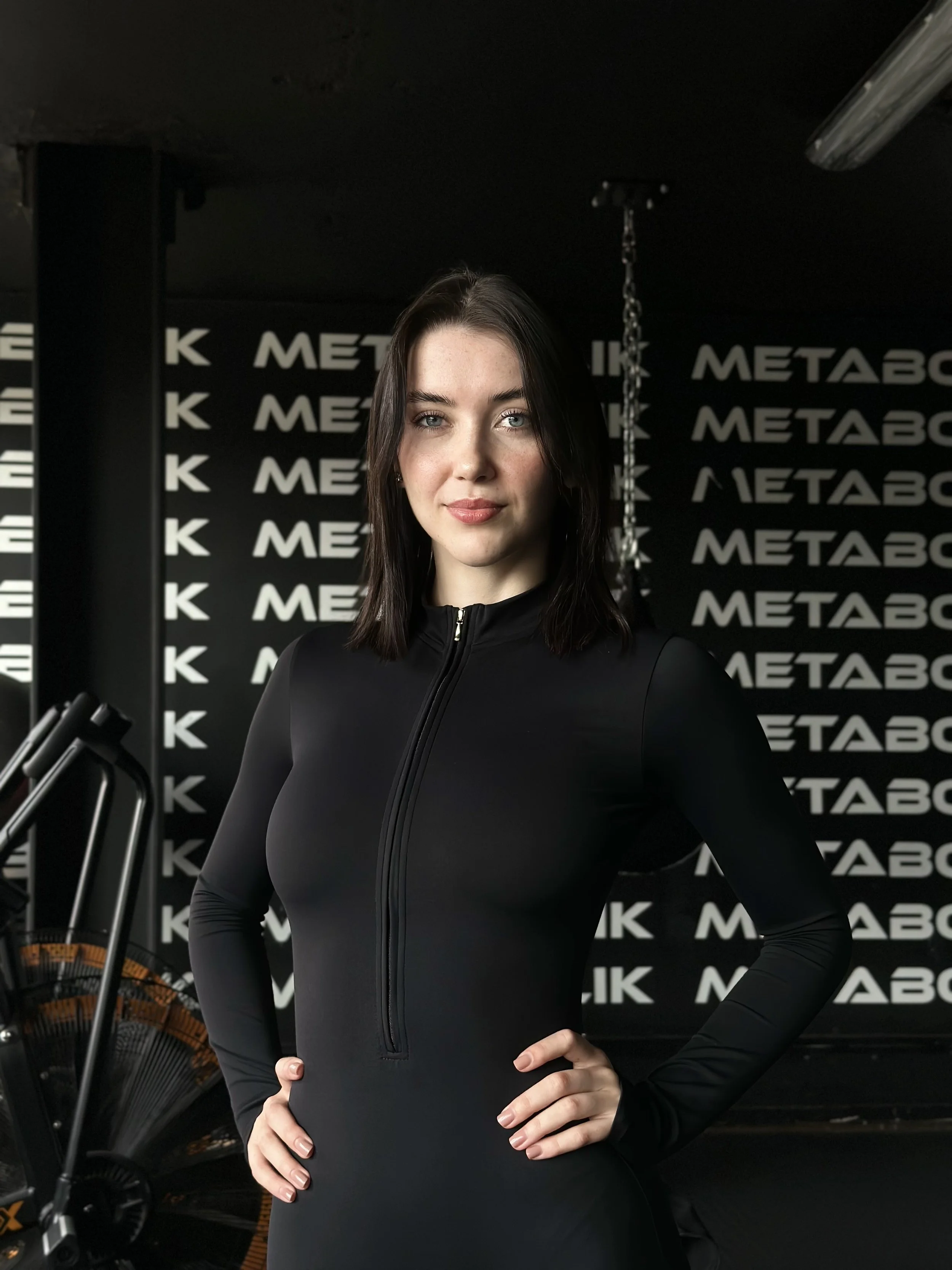 A young woman with dark hair and fair skin in a black athletic outfit standing with hands on hips in a gym.