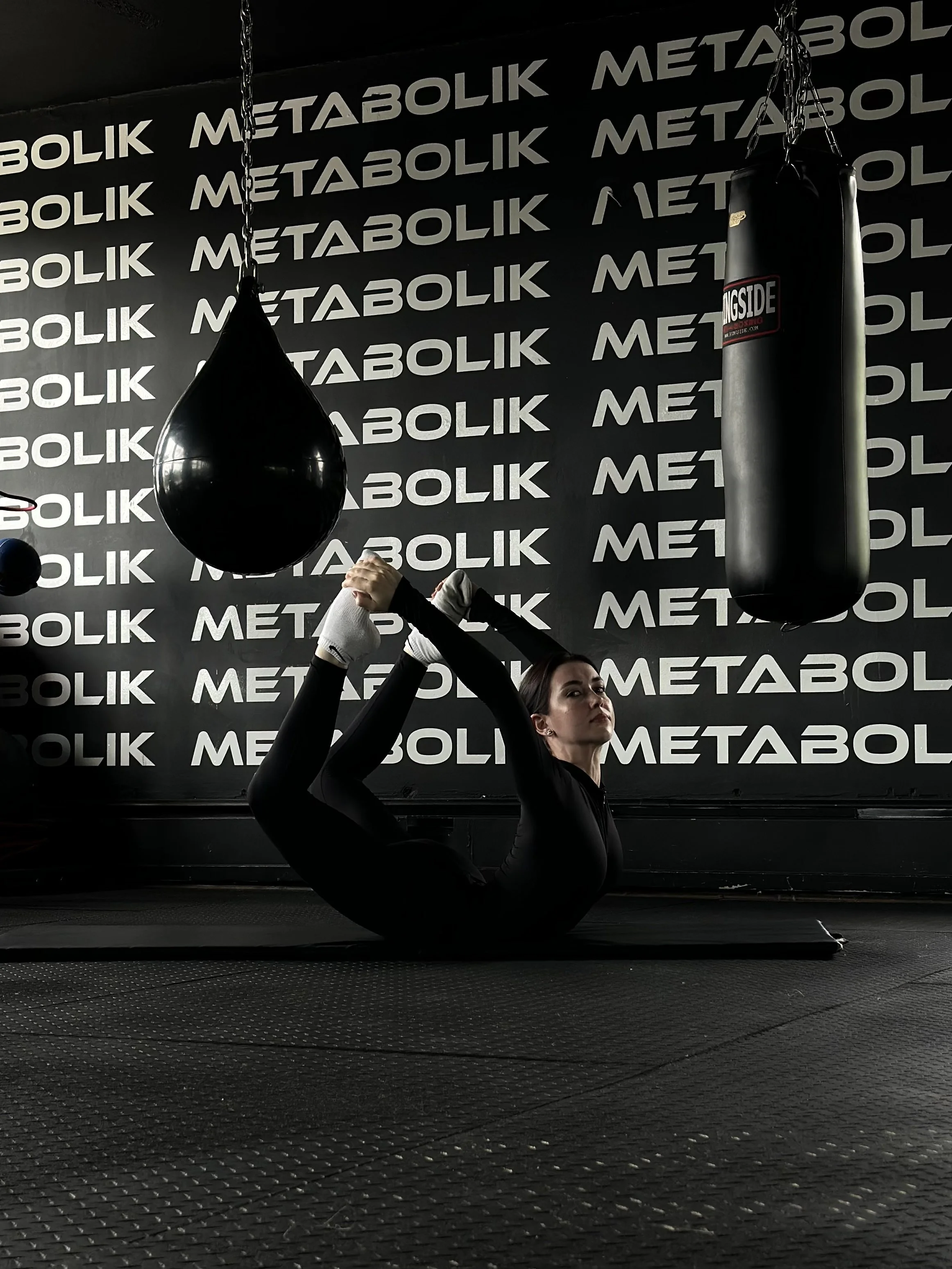 A woman in black workout attire practicing yoga or stretching on a black mat inside a gym with black walls, which have the word 'METABOLIK' repeated in white.