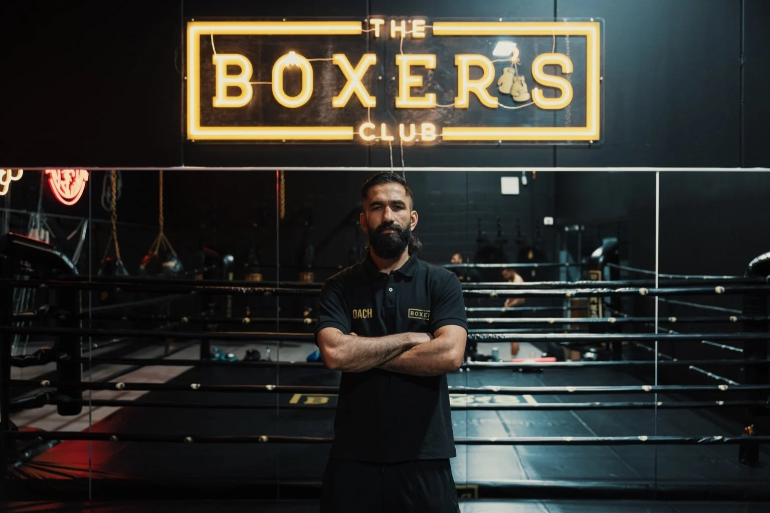 A bearded man with crossed arms standing in front of a boxing ring inside a gym, with a neon sign above reading 'The Boxers Club'