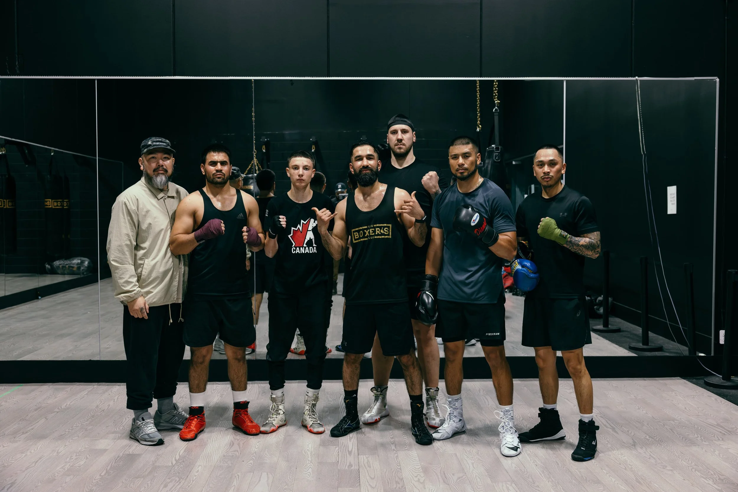 A group of seven men, some wearing boxing gloves, standing inside a boxing gym with black walls and a mirror behind them. They are posing confidently for the photo.