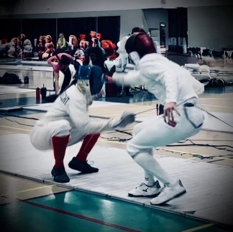 Two fencers in white uniforms and masks engaged in a match at an indoor fencing competition, with spectators and other fencers in the background.