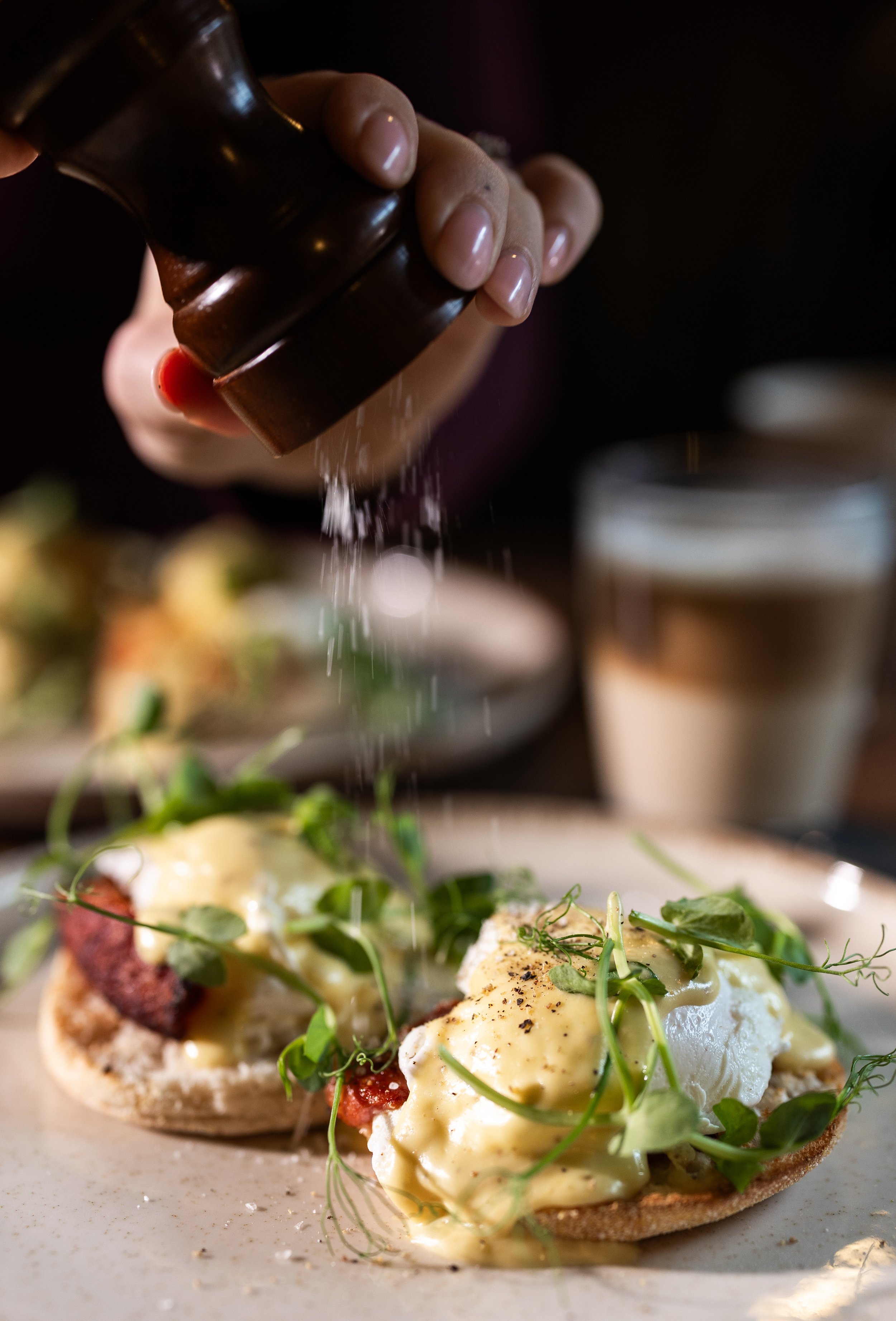 A person sprinkles salt onto eggs Benedict served on toasted English muffins garnished with microgreens, with a glass of beer in the background.