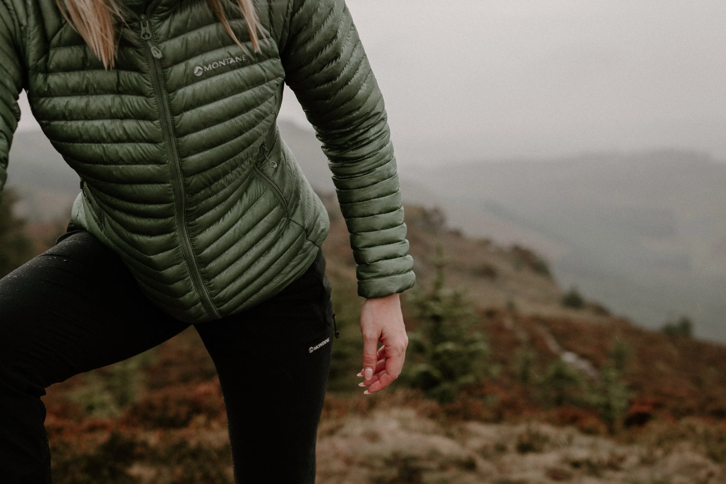 Person in a green Montana quilted jacket and black pants standing on a misty mountain trail with small trees and a body of water in the background.