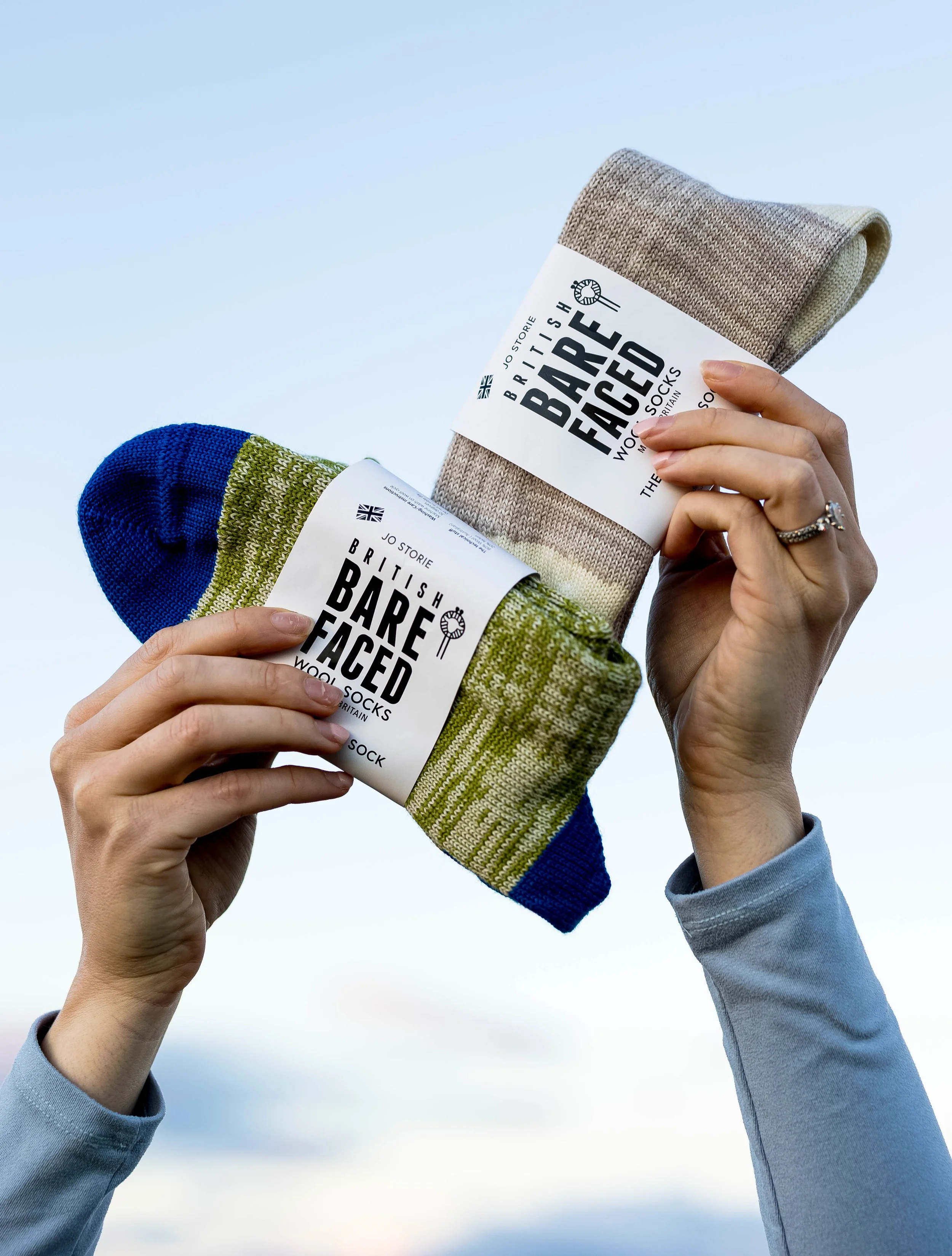 Two people holding packaged socks called 'Bare Faced', with a blue and green sock in one hand and a beige sock in the other, against a clear sky background.