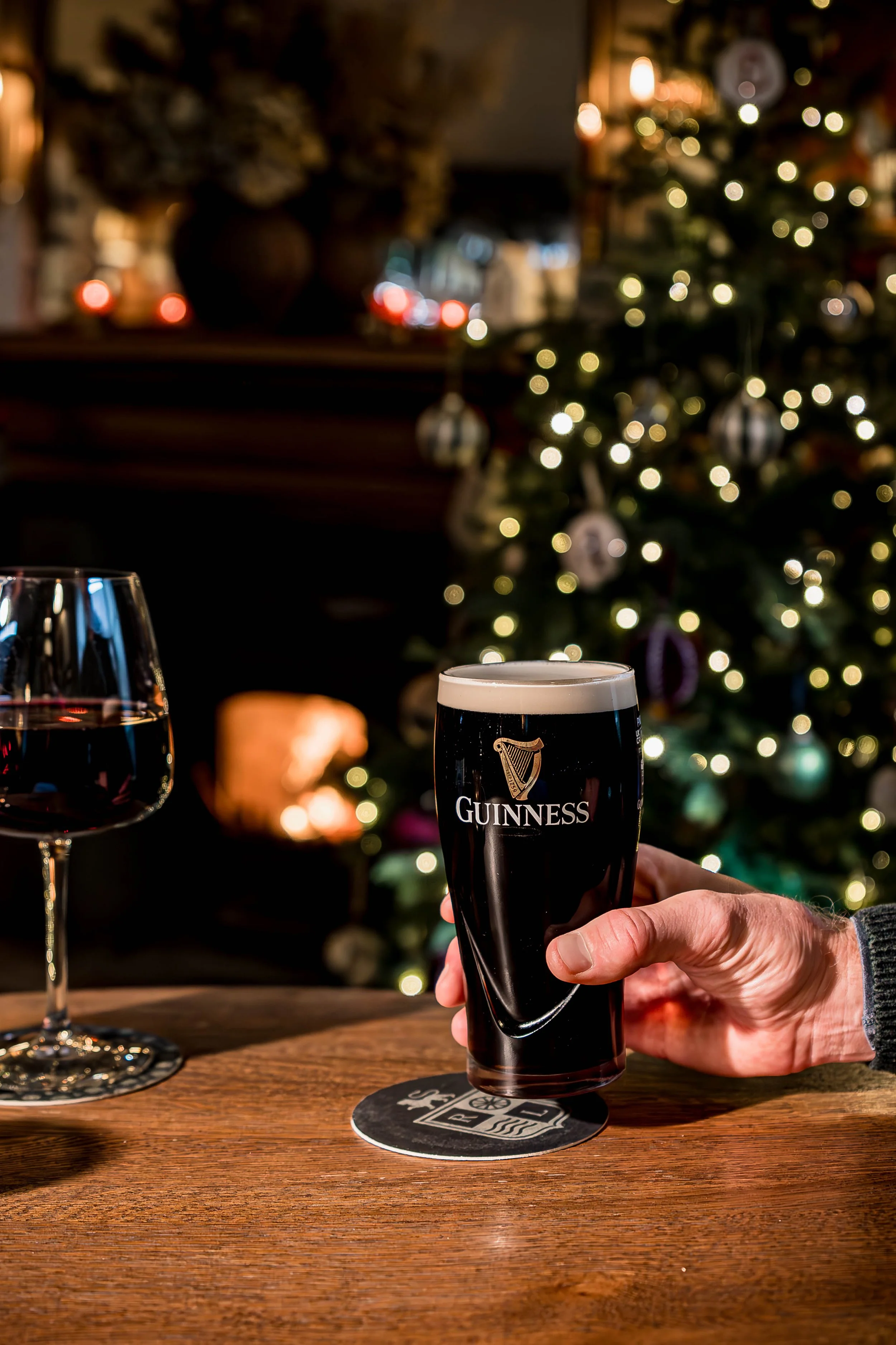 A person holding a pint of Guinness beer at a Christmas-themed setting with a decorated Christmas tree and warm lighting in the background.