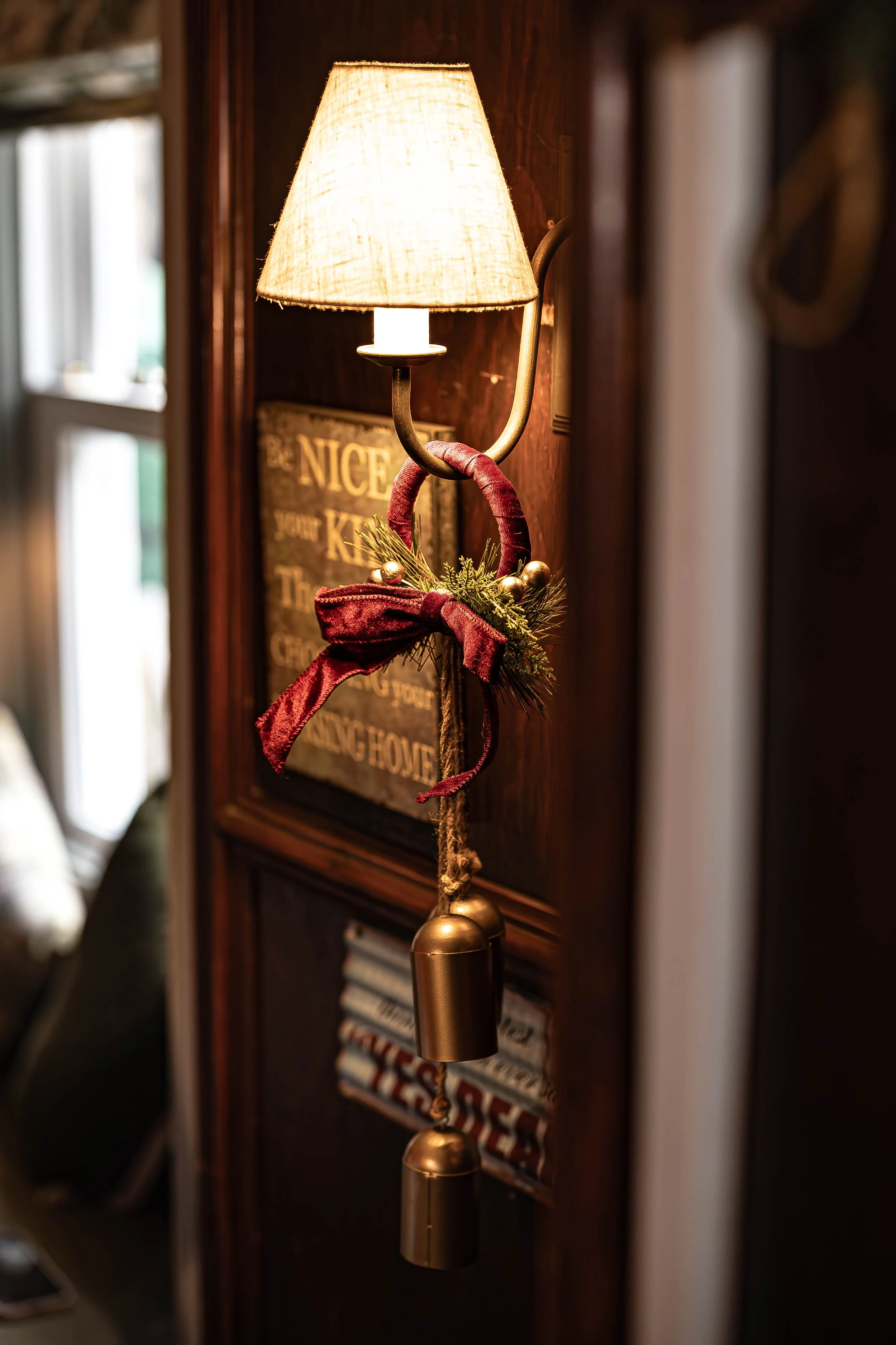 A wall-mounted lamp with a decorated red velvet bow and greenery, featuring pine branches and gold ornaments, hanging on a dark wooden wall.